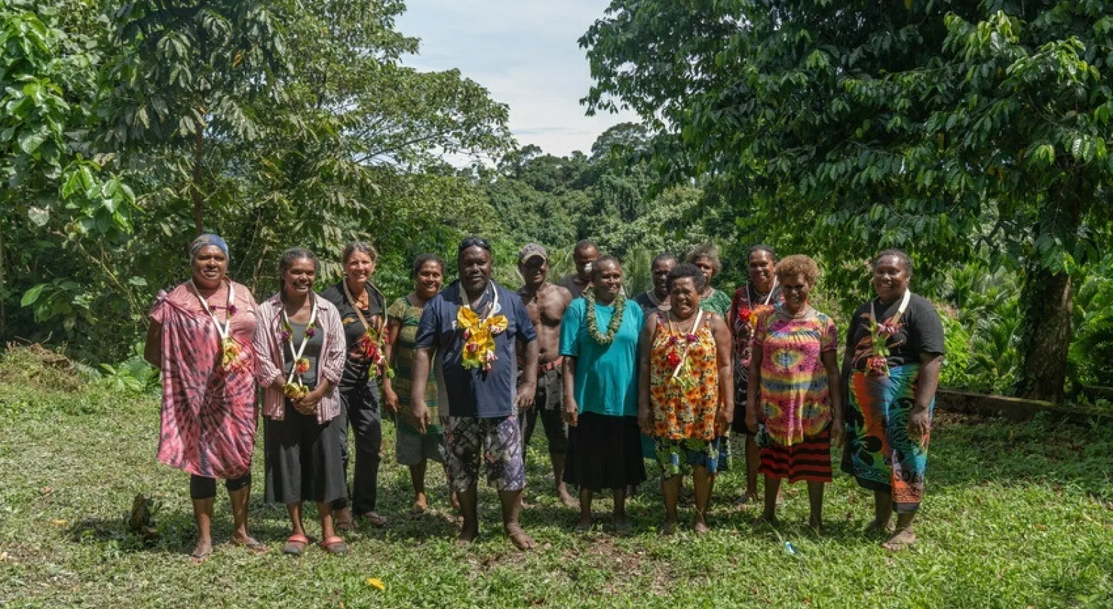 A group of 14 people in a variety of colours are standing together in a lush, outdoor setting.