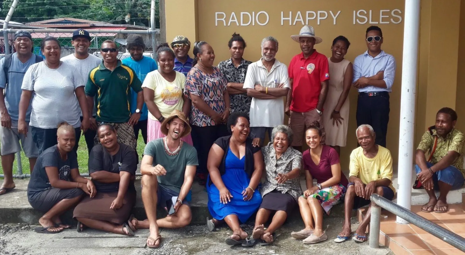 A group of people gathered outside a building, with a sign that says 'S.I.B.C Radio Happy Isles' behind them. The people in the photos are smiling and laughing.