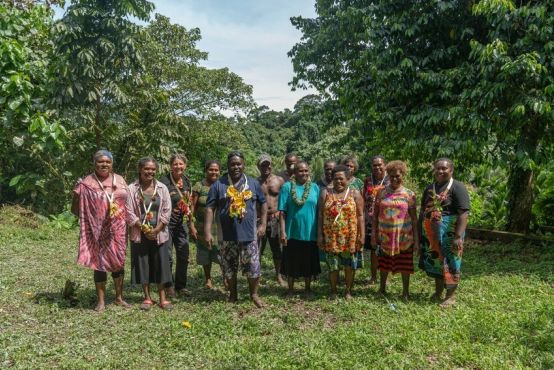 A group of 14 people in a variety of colours are standing together in a lush, outdoor setting.