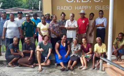 A group of people gathered outside a building, with a sign that says 'S.I.B.C Radio Happy Isles' behind them. The people in the photos are smiling and laughing.