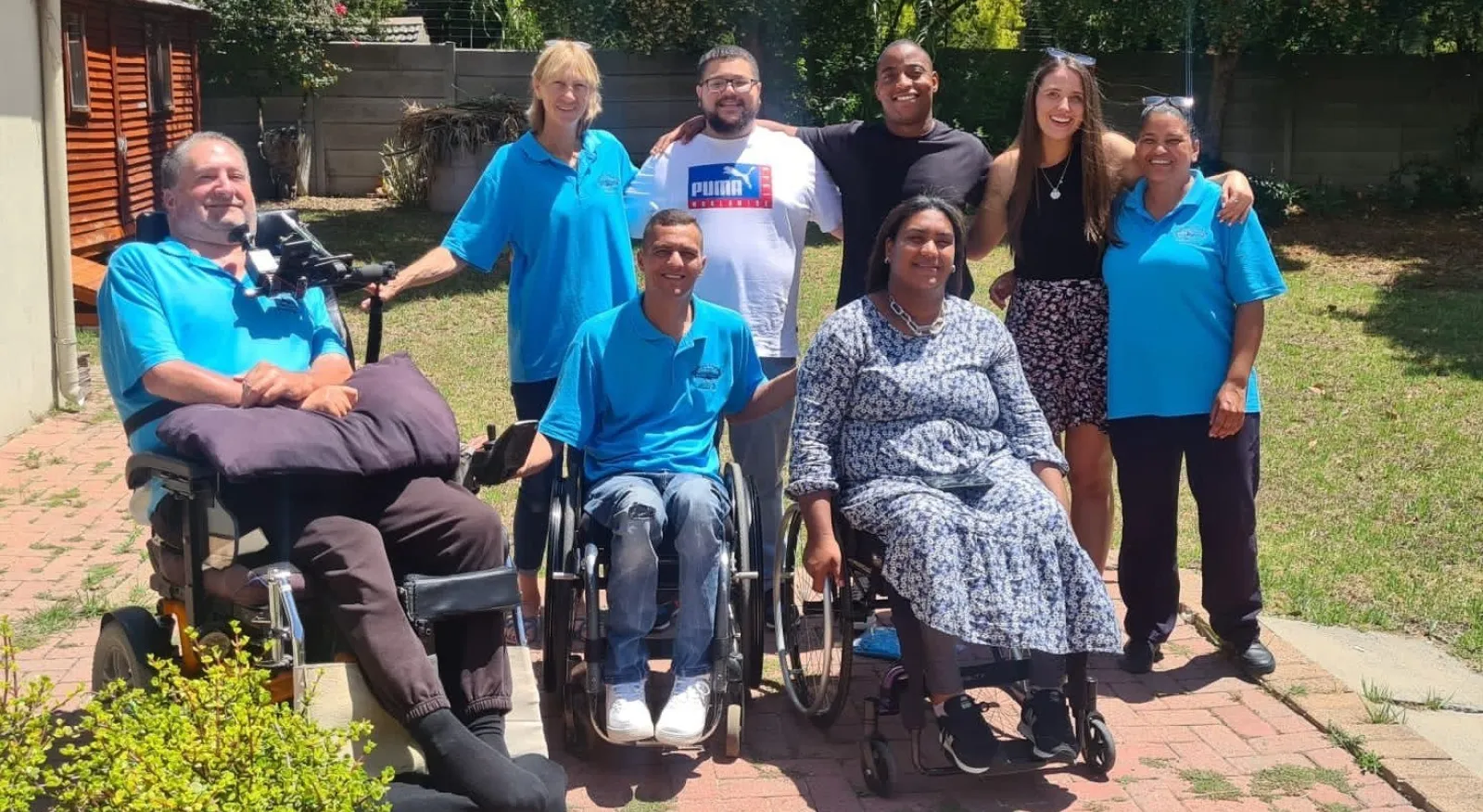 Eight people smiling together in an outdoor setting; three people are in wheelchairs and four people are standing and have arms around each other.