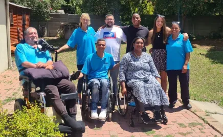 Eight people smiling together in an outdoor setting; three people are in wheelchairs and four people are standing and have arms around each other.