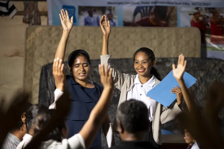 Two people standing at the front of a classroom, each with a hand raised, in front of students with their hands raised.