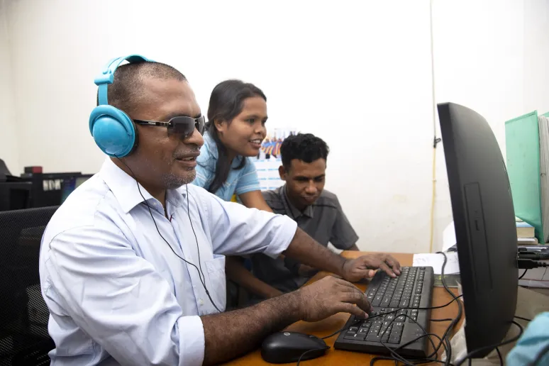 Three people are huddled around a computer, facing the desktop screen. The person in the foreground has blue headphones and is operating the keyboard.