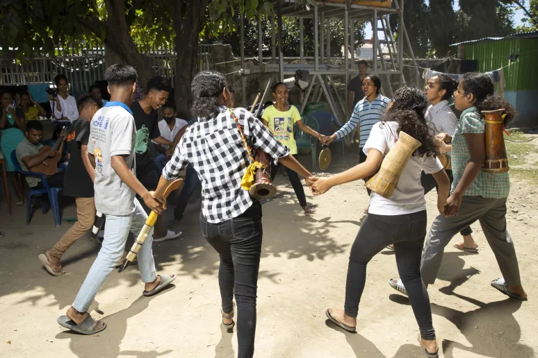 A group of people are linking hands in a circle and dancing in an outdoor setting.
