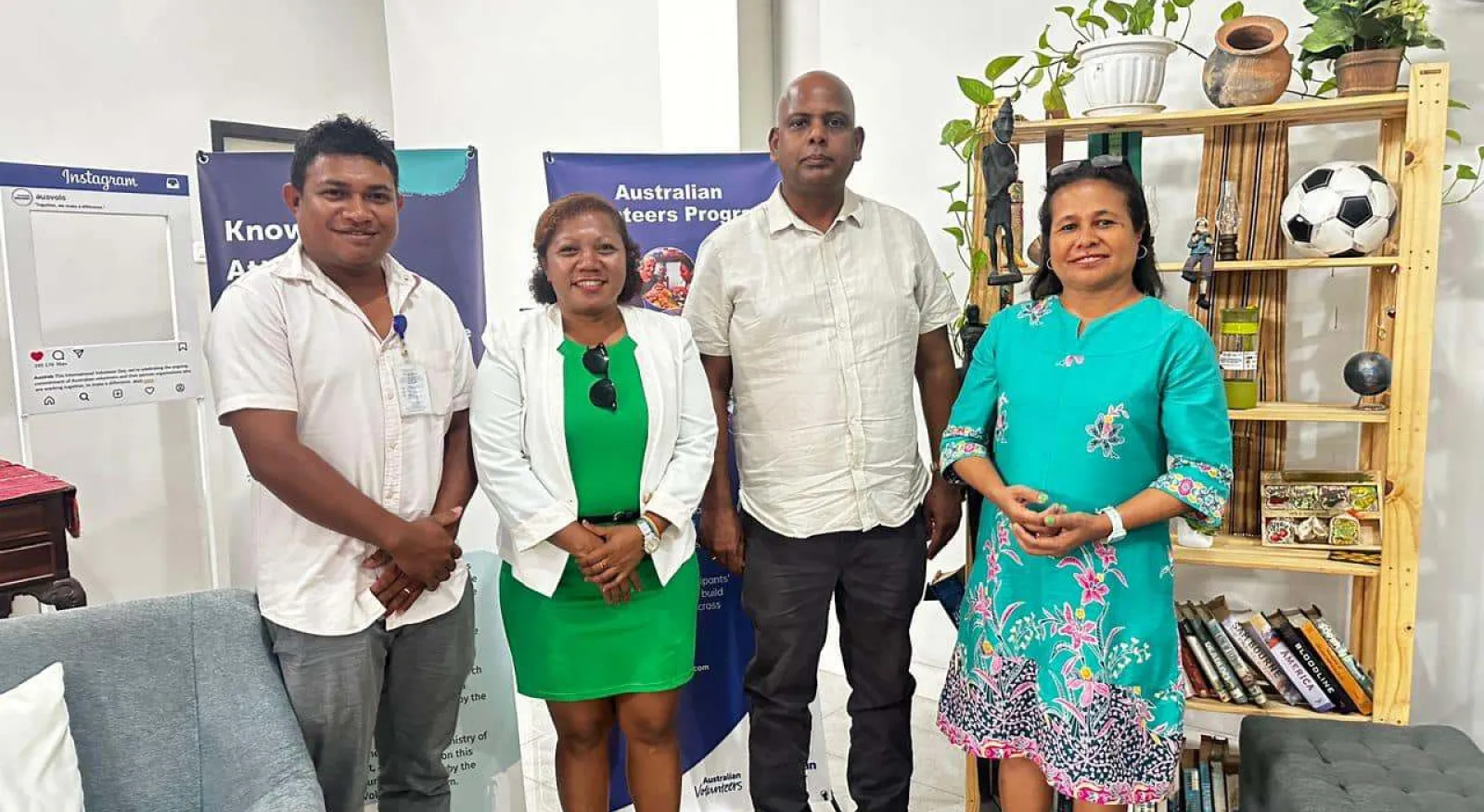 Four people stand in a line facing the camera and smiling. Three have their hands folded in front of them, one has their hands by their sides. In the background are banners, furniture and artifacts typical of an office or shared work space.