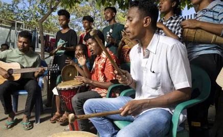 A colourful group of people - some sitting and some standing - are playing musical instruments together under the shade of a tree.