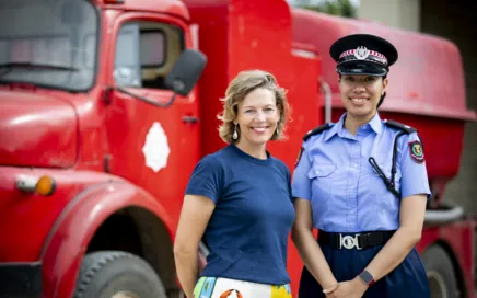 Two women stand in front of a red fire truck and smile at the camera. One woman is wearing casual clothing, the other wears the fire services uniform including hat, collared shirt with badges and radio.