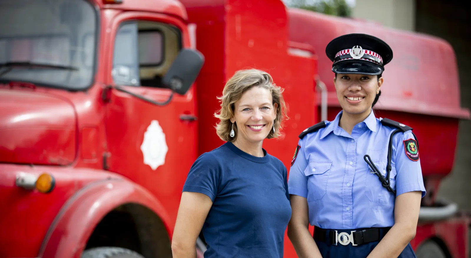 Two women stand in front of a red fire truck and smile at the camera. One woman is wearing casual clothing, the other wears the fire services uniform including hat, collared shirt with badges and radio.