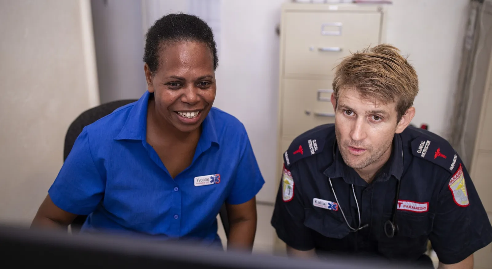 Two people are looking at a computer screen. The person on the left is smiling. Both are wearing blue uniforms.