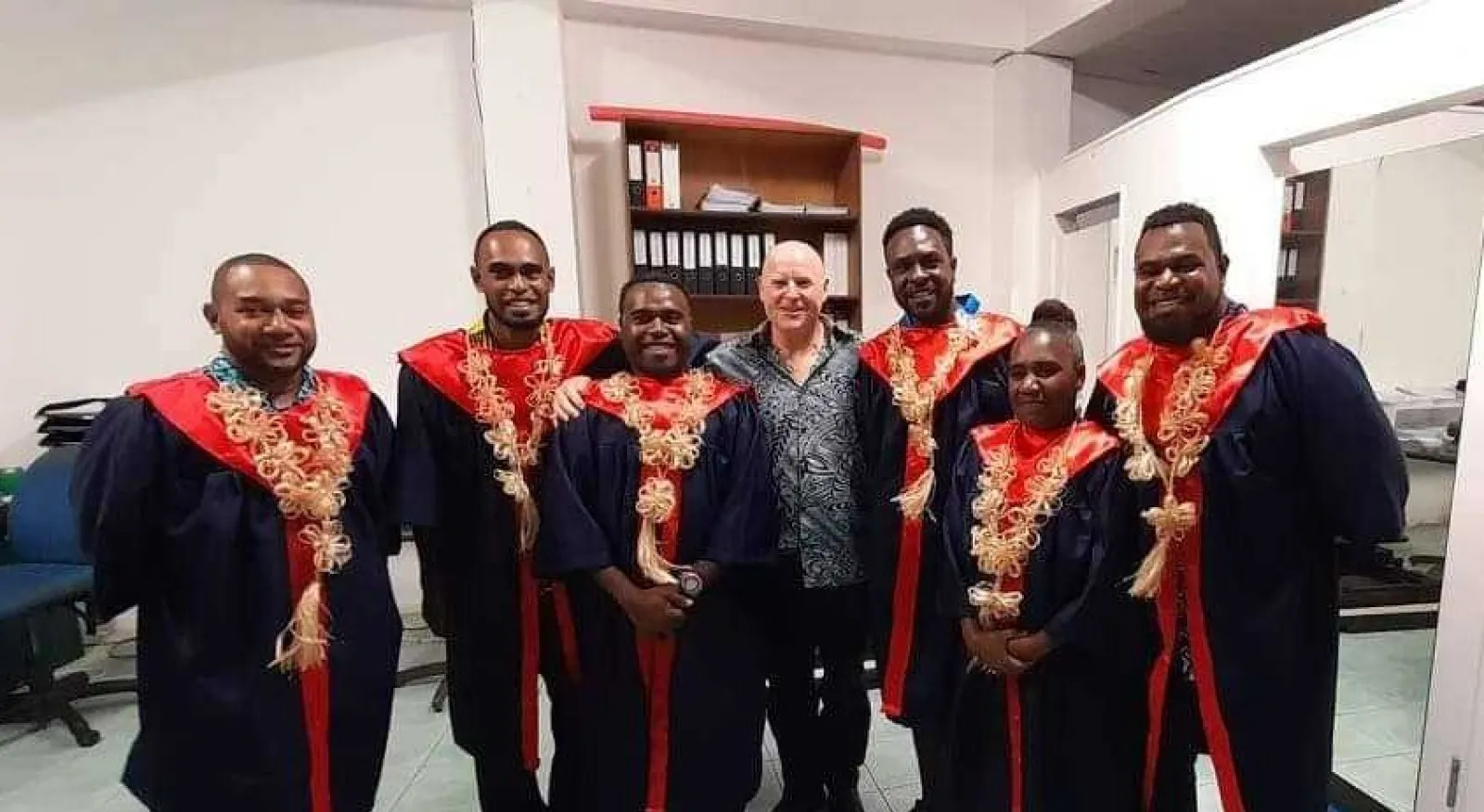 Six students in academic gowns stand next to each other and smile into the camera. Standing in the middle, with three students on either side, is volunteer Wayne Barnes who also smiles into the camera.