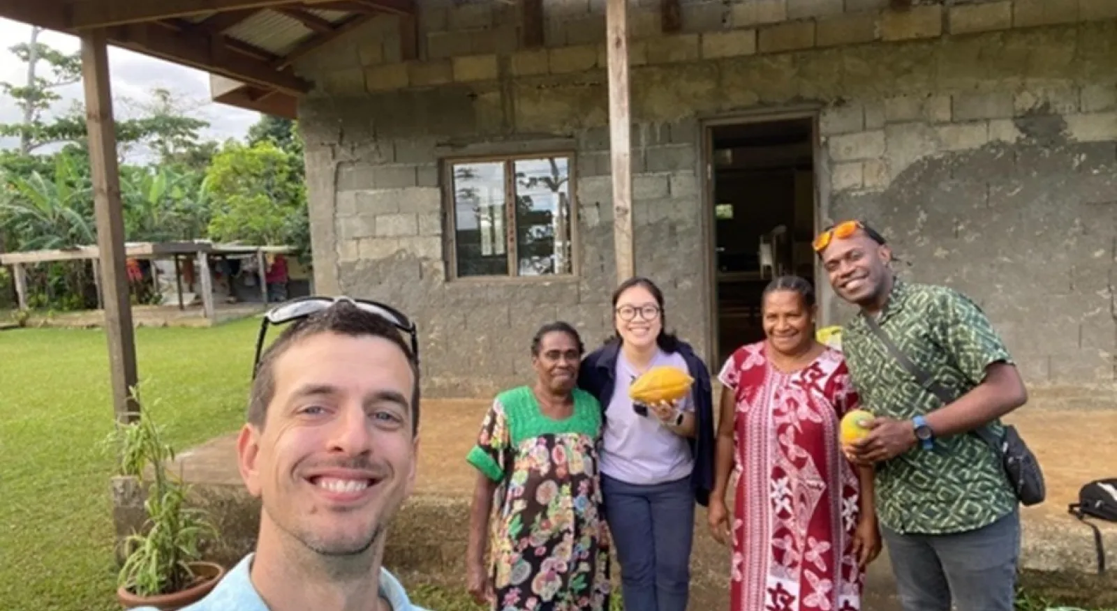 A 'selfie'; in the foreground is the person taking the photo, and in the background four people stand side-by-side outside a building surrounded by grass and vegetation in the far distance. Everyone is smiling into the camera.