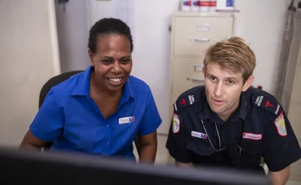 Two people are looking at a computer screen. The person on the left is smiling. Both are wearing blue uniforms.
