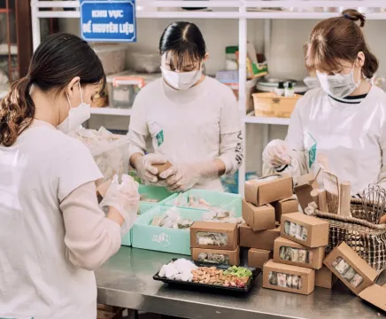 Three women are wearing white chef shirts and face masks, all working on cookies in an industrial kitchen.