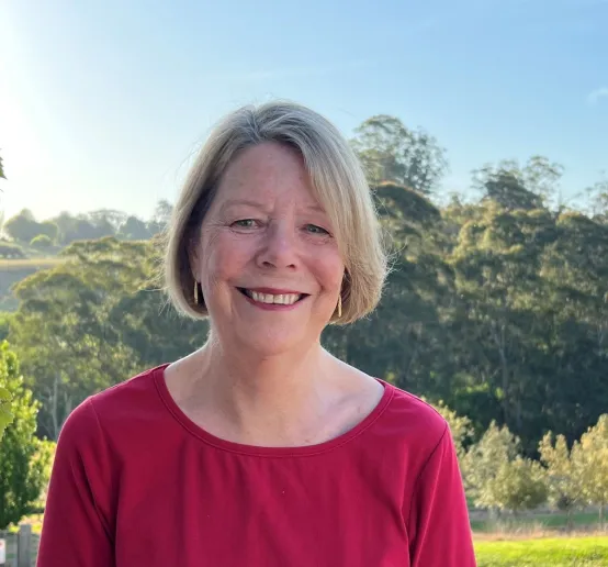 A woman with short blonde hair and a red shirt smiles at the camera. Behind her is a scenic background of of gum trees and a rolling hill.