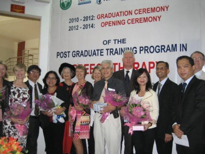 A group of 12 people stand in front of a sign that is partially visible and says "Graduation Ceremony of the Post Graduate Training Program." Some people have flowers and graduation regalia. Everyone is smiling at the camera.