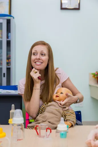 A red-headed woman is smiling at someone off-camera. She holds a baby doll in her lap, supporting his head.