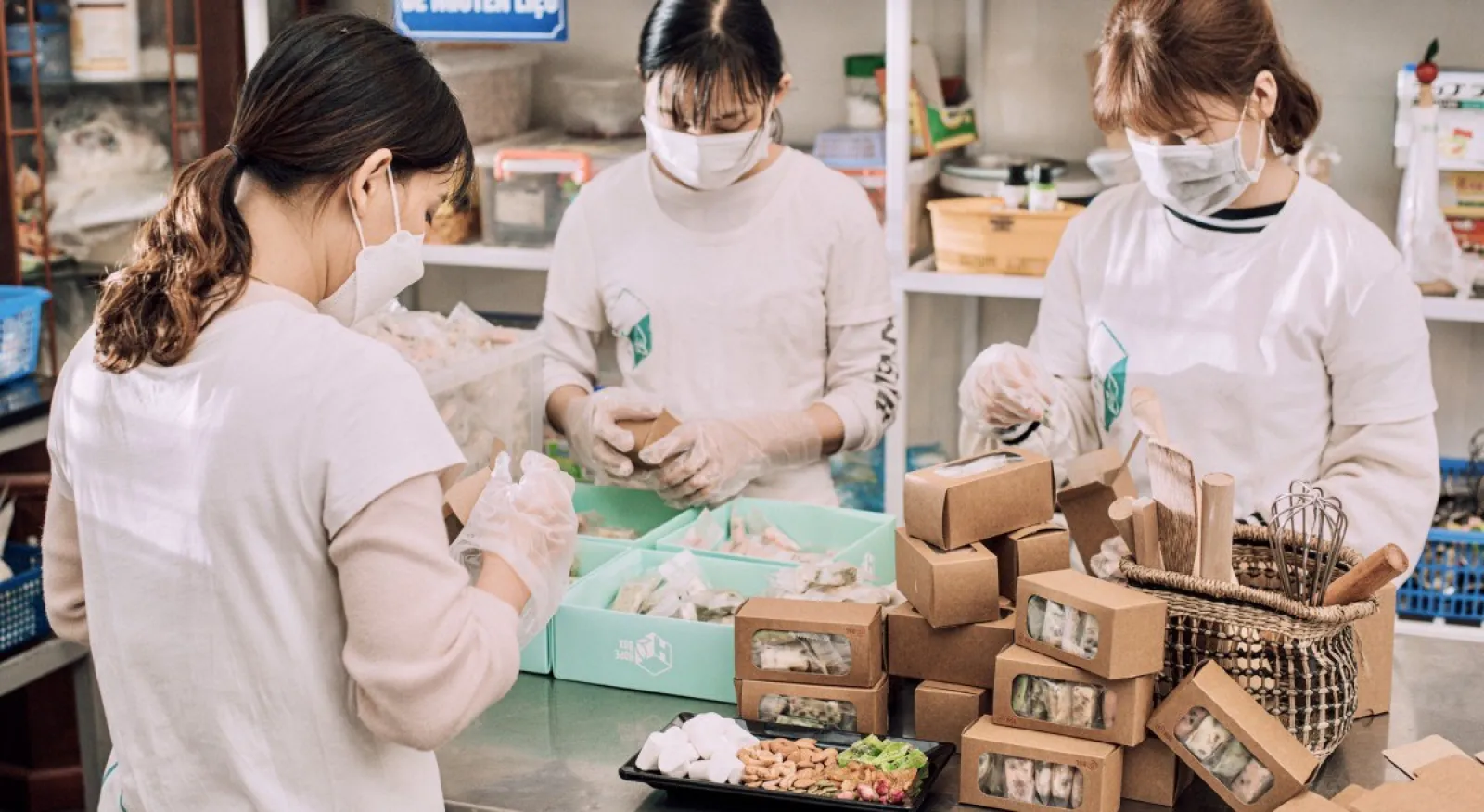 Three women are wearing white chef shirts and face masks, all working on cookies in an industrial kitchen.