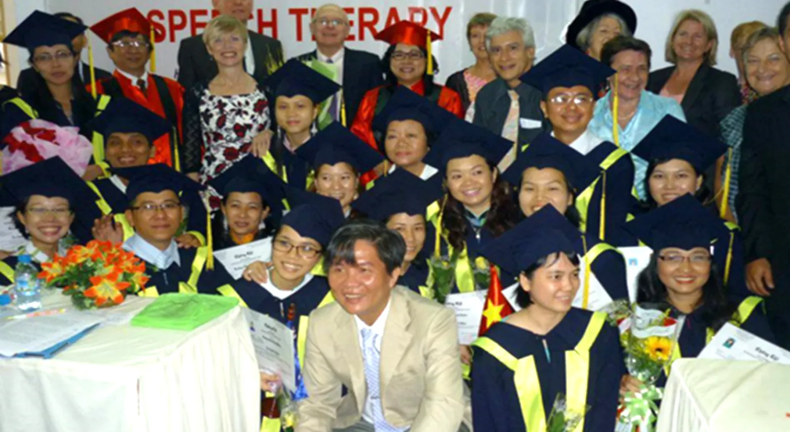 A group of individuals wearing graduation gowns and caps, celebrating their academic achievements. There is a banner behind them saying 'Post graduate training program in speech therapy'