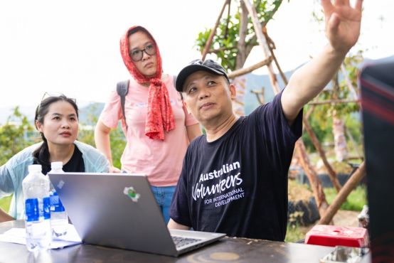 A man and woman sitting at a table, working on their laptop, while another women is standing and watching over.