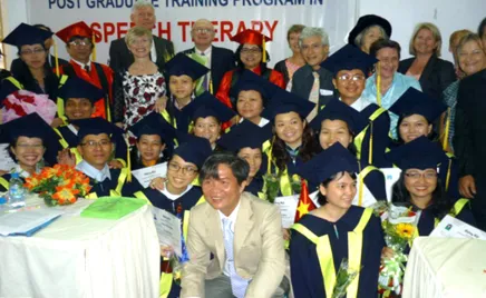 A group of individuals wearing graduation gowns and caps, celebrating their academic achievements. There is a banner behind them saying 'Post graduate training program in speech therapy'