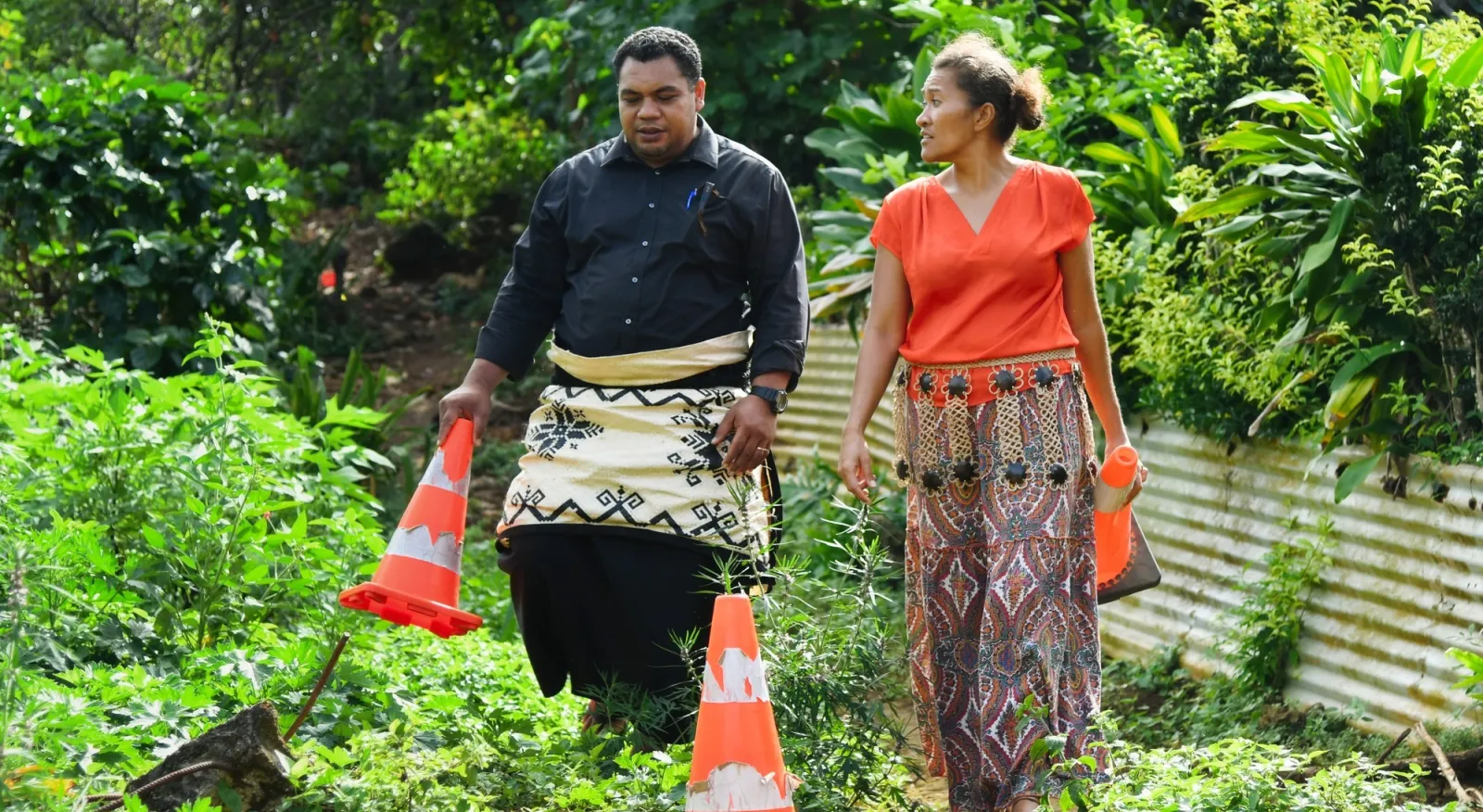Two people in traditional Tongan dress are laying cones down on a very green, vegetated road. They are mid-discussion and not looking at the camera.