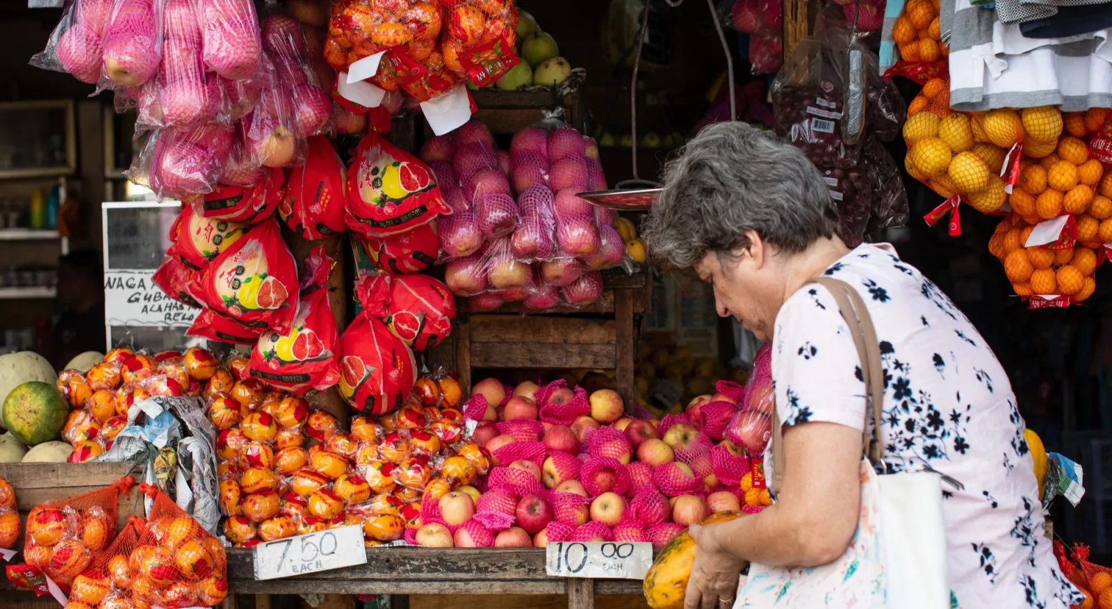 A woman stands in front of a full market stall, her back is to the camera and she inspects the fruit on the shelves.