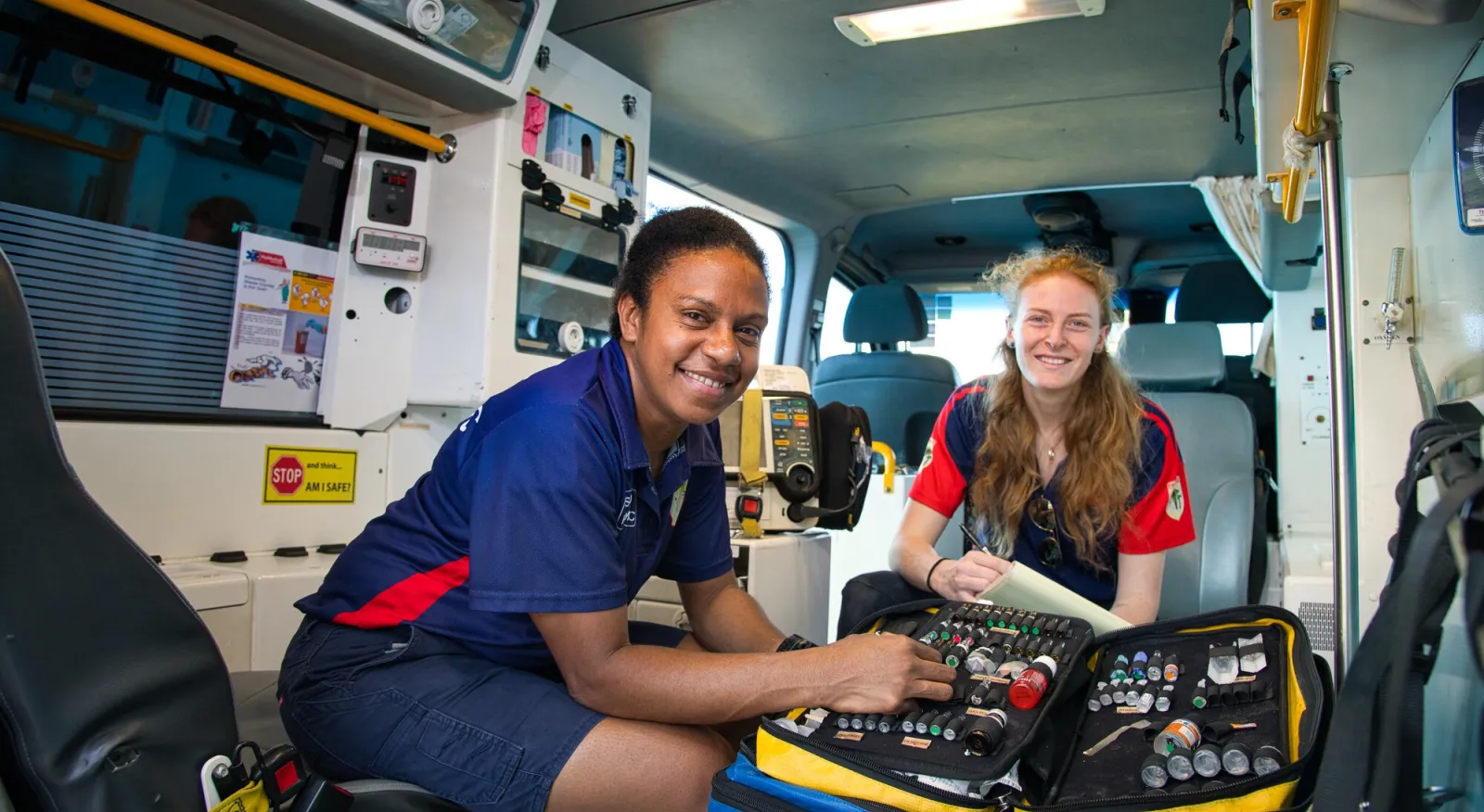 Two women sit in the back of an ambulance, they are paramedics wearing the uniform. They are smiling to the camera and a medic bag lays open between them.