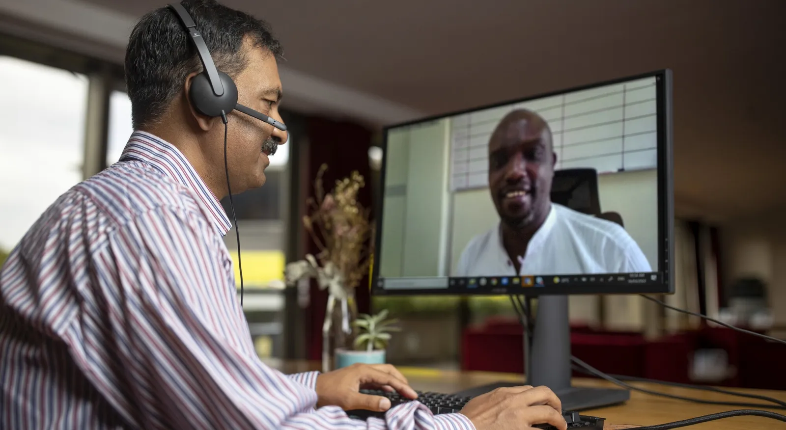 Two men are on a Zoom call together. One man is in the room, with his side to the camera. On his screen is another smiling man.