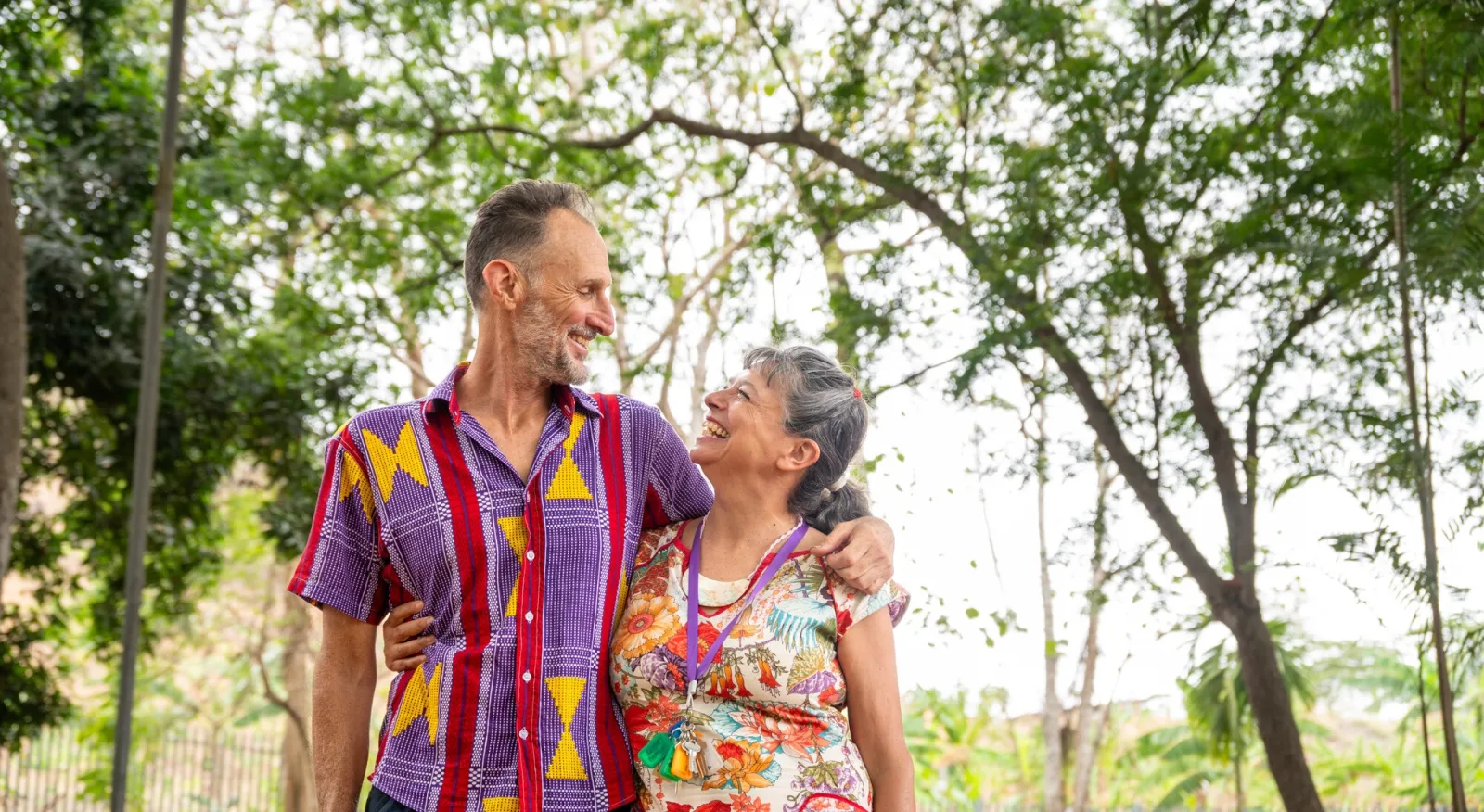 A man and a woman stand in front of trees, they have their arms wrapped around each other's shoulder and are smiling at each other.