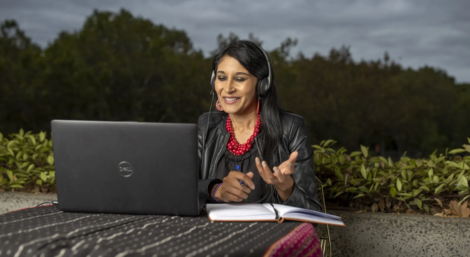 A woman sits at a laptop, wearing a black jacket and tanktop as well as a bright red necklace. She is outside, and has headphones on. It looks like she is mid conversation with someone on screen.