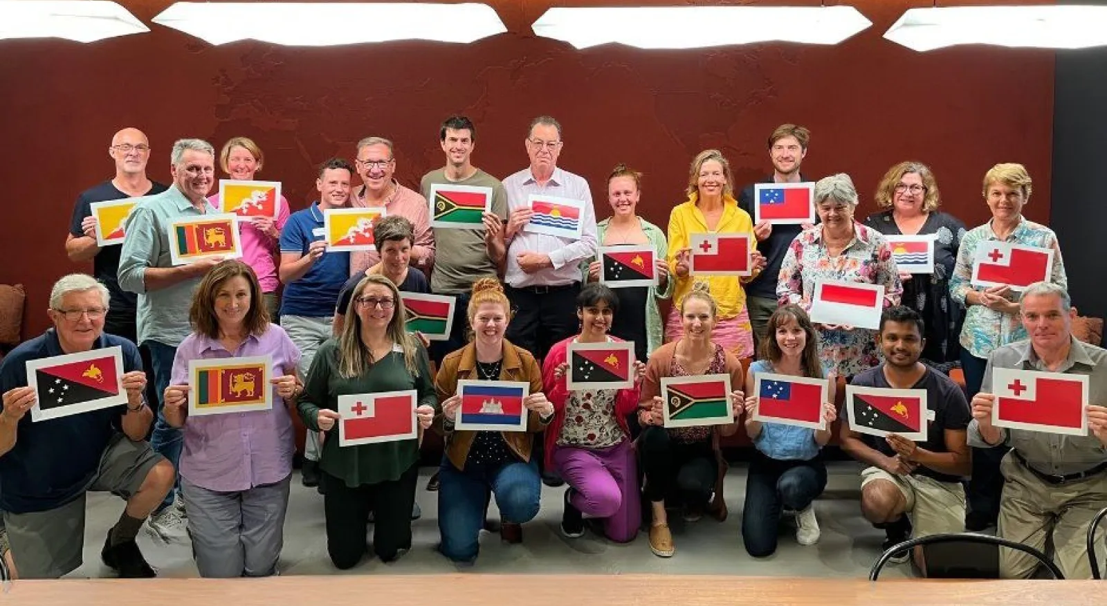 A large group of people are gathered for a group phto against a bright red wall. They all hold paper copies of flags where they will be volunteering.