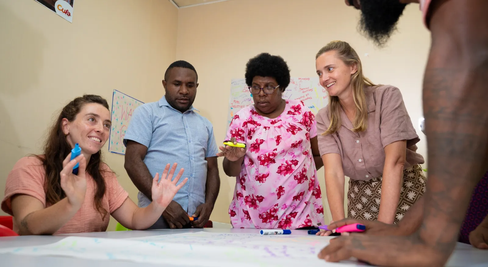 A group of five people sit around the table holding multi-coloured markets. They are mid-conversation and drawing on large pieces of paper.