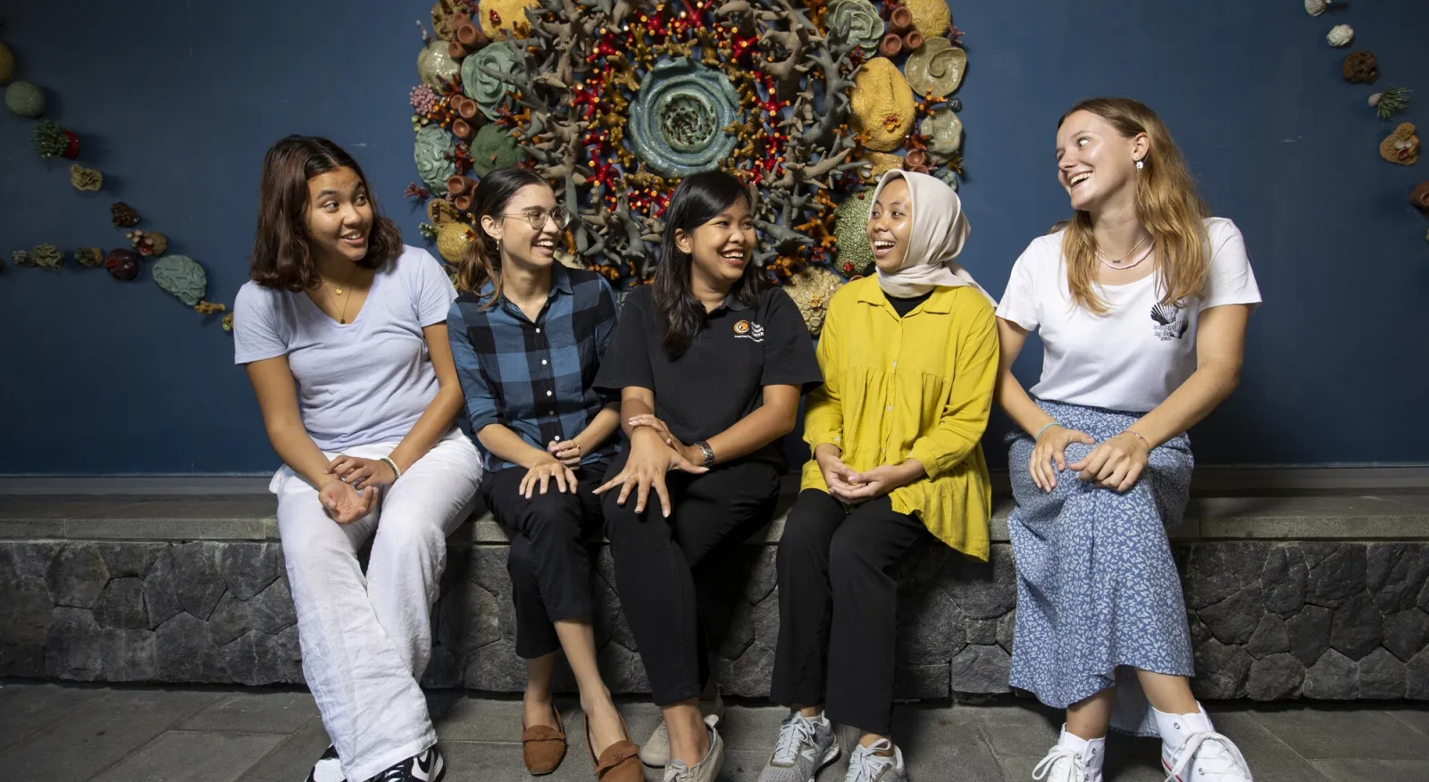 Five women sit in front of a sculpture, they are all laughing and looking at each other.