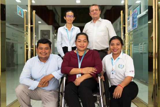 A group of people are posed in the front of a gold doorway, all in professional workplace dress. One woman is in a wheelchair, with another person kneeling on either side of her. Two people are standing behind them. All are smiling at the camera.