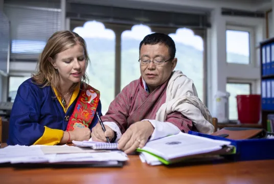 Woman and man sit at desk working on documents together, both wear traditional Bhutanese dress