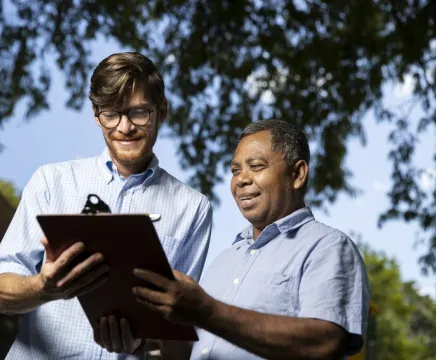 Two men in light blue shirts stand outside and look at a clipboard