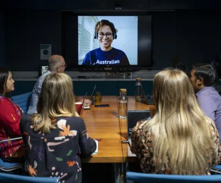 A group of people sit around a board table looking at a screen with a woman in a blue Australian Volunteers t-shirt on it