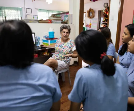 A lady in a colourful top with short grey hair sits talking to a group of students