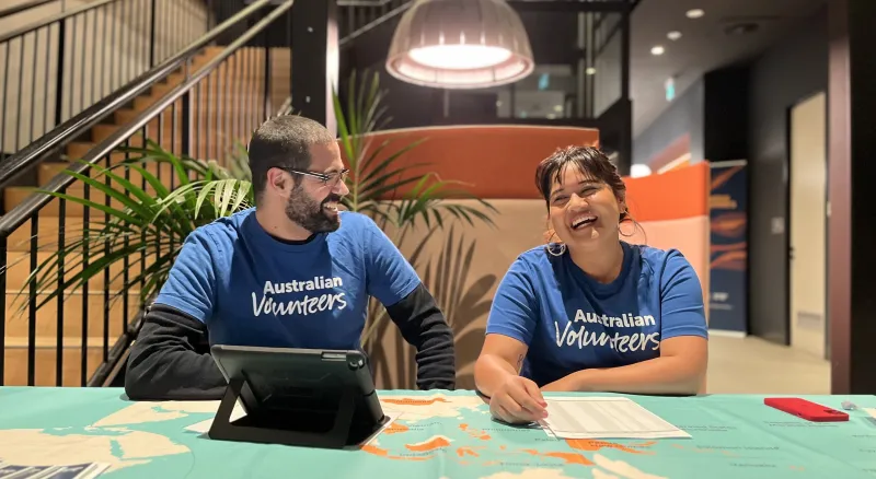 A man and a lady wearing Australian volunteers t-shirts sit at a table laughing