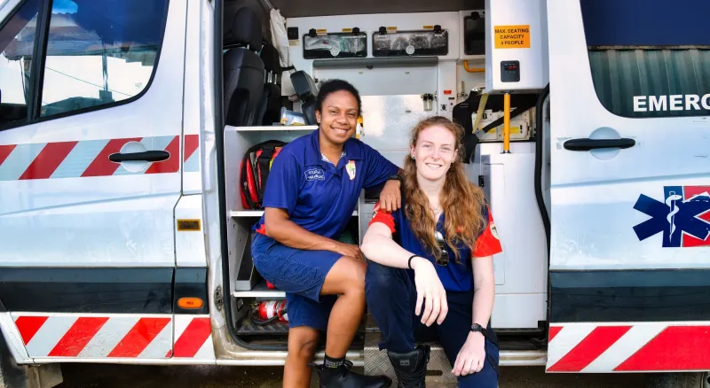 Two women stand in front of an open ambulance, they are in two different medical uniforms