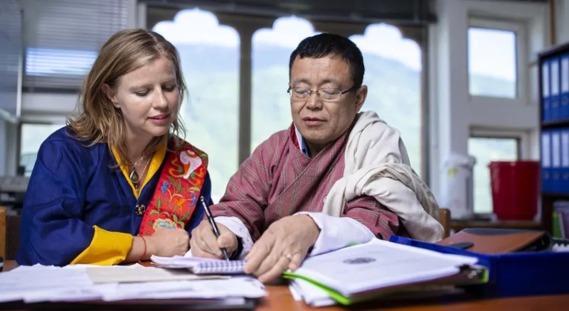 Woman and man sit at desk working on documents together, both wear traditional Bhutanese dress