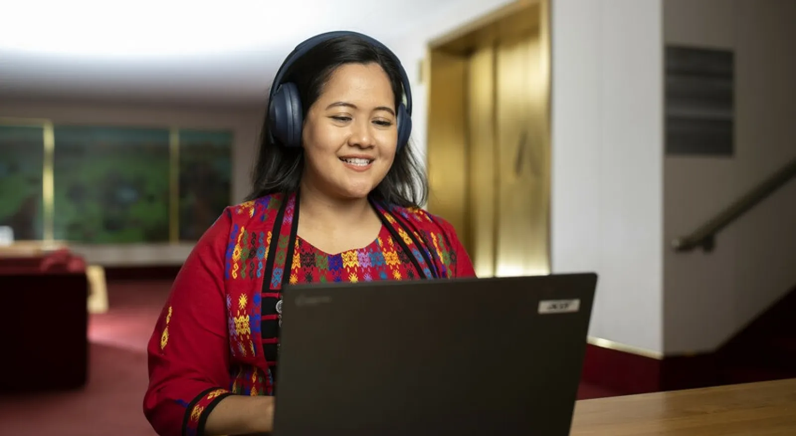 A lady in a red embroidered top sits inside, she is on a laptop and has headphones on