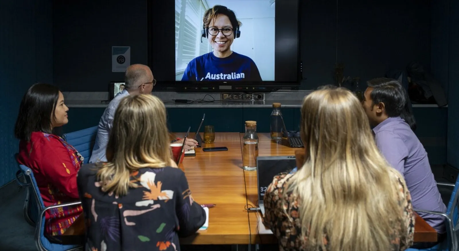 A group of people sit around a board table looking at a screen with a woman in a blue Australian Volunteers t-shirt on it