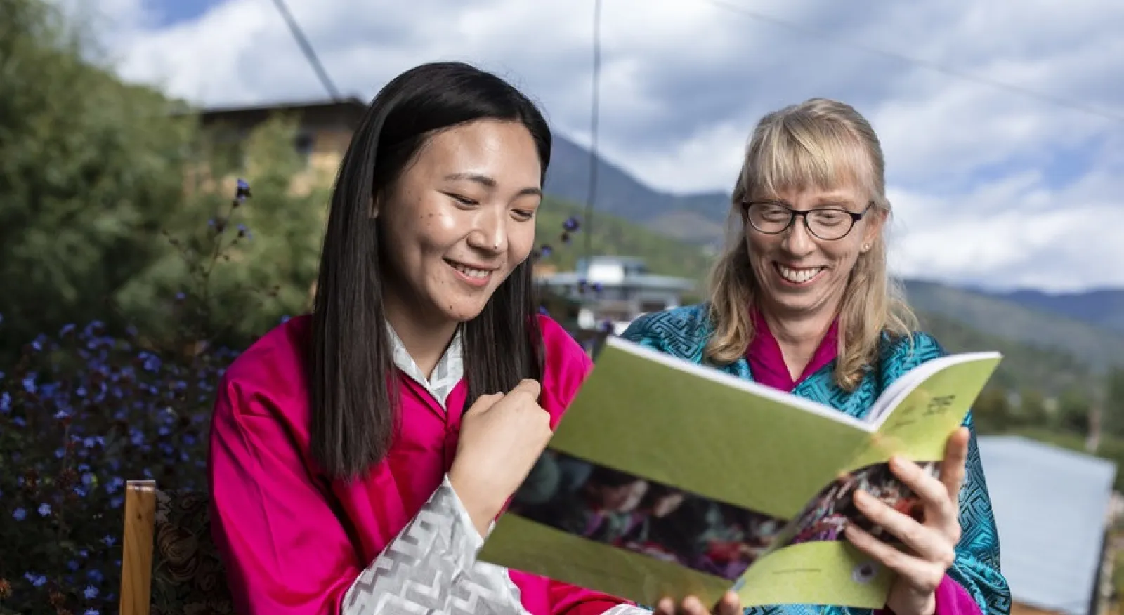 A dark haired lady in a bright pink top reads a book with a light haired lady in a aqua top.. They are in front of a background of Bhutanese mountains.