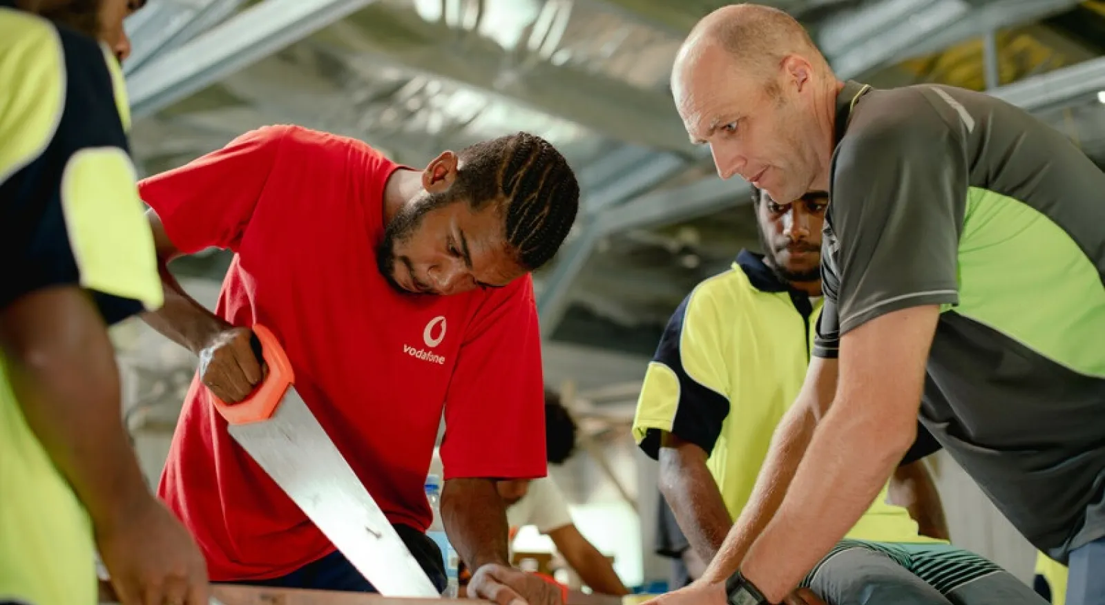 A man in a red shirt uses a hand-saw, he is supervised by a bald man in a grey and yellow shirt. Two others watch on.