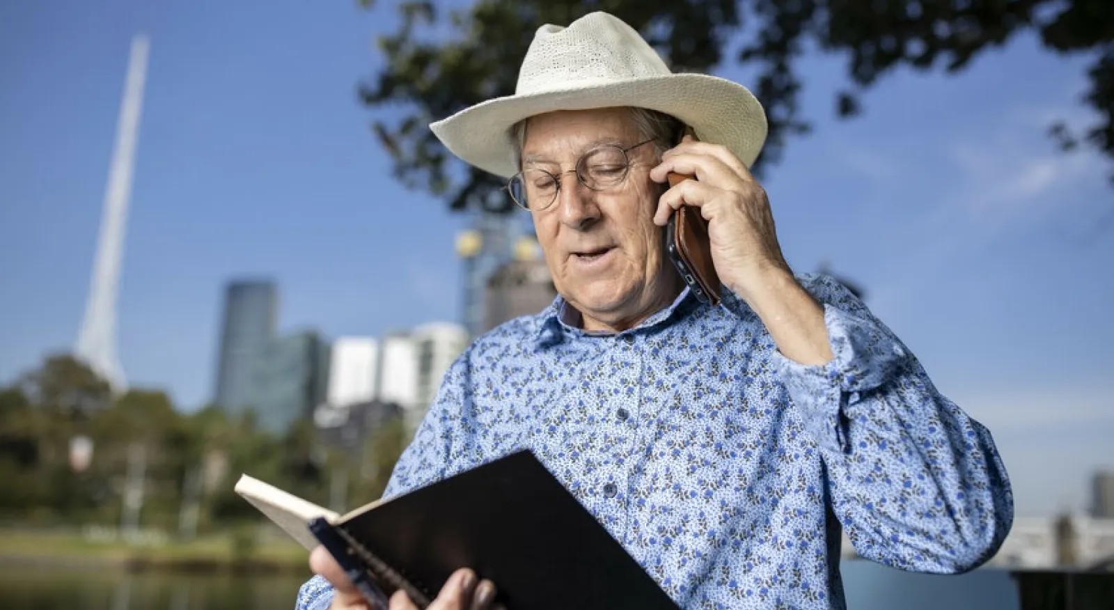 An older man in a blue long-sleeved shirt and wide-brimmed hat is on the phone, looking at a book. In the background is the skyline of Melbourne
