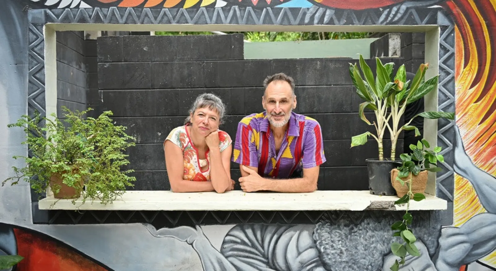 two people lean on a ledge in a window, framed by colourful artwork