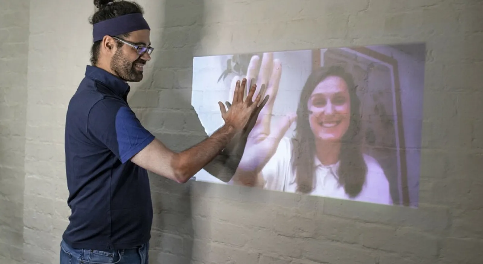 a person looks at a projection on a wall and holds their hand up to match the hand of a person in the projection. Both smile