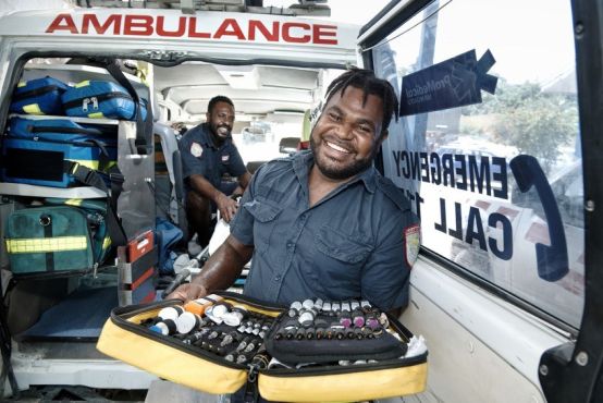 A smiling man in a blue uniform stands in front of an ambulance holding open a yellow case full of medical equipment, in the background is an open ambulance which another man in uniform is sitting in